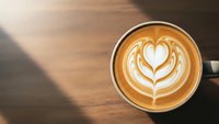 Coffee cup with heart latte art on wooden table with natural sunlight creating warm atmosphere for cafe morning routine and beverage enjoyment.