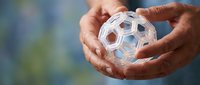 Hands gently holding transparent geometric sphere with hexagonal pattern against soft blue background. Science, chemistry, molecular structure concept.