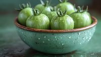 Fresh green unripe tomatoes with water droplets in a rustic turquoise ceramic bowl on dark surface. Perfect for farm-to-table food concepts.