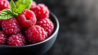Fresh ripe raspberries with green mint leaf in black ceramic bowl on dark background, macro shot with selective focus and natural light.