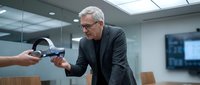 Senior male professional with white hair and glasses, wearing dark blazer, examining virtual reality headset in modern office conference room with glass walls and wooden furniture.