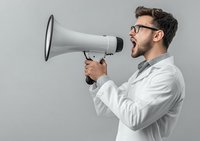 Young businessman in white shirt and glasses shouting into megaphone for marketing communication and announcement campaigns on gray background.