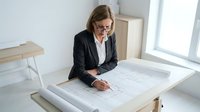 Professional businesswoman architect reviewing construction blueprints at modern office desk, focused on technical drawings and building plans.