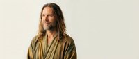 Middle-aged Caucasian man with long brown hair and beard wearing olive green robe, smiling peacefully against neutral background for wellness content.