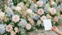 Unrecognizable person holding blank white card in front of blooming garden with pink roses and white flowers for greeting, invitation, or promotional messaging.
