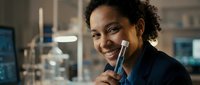 Young African American woman smiling while holding makeup brush in modern beauty salon or cosmetics store with professional lighting and mirrors.