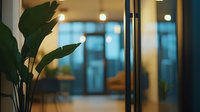 Modern office interior with large green plant leaves in foreground, blurred glass doors and warm lighting creating cozy professional atmosphere. Perfect for business decor.