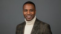 Professional African American businessman in brown suit and turtleneck smiling confidently against gray background for corporate headshot photography.