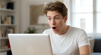 Young Caucasian man with surprised shocked expression looking at laptop screen in modern home office, reacting to unexpected news or information online.