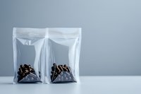 Two transparent plastic zip lock bags containing dark roasted coffee beans on light gray background, minimalist composition for food packaging and retail presentation.