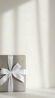 Elegant gray gift box with white satin ribbon bow on a light background with soft window shadows for holiday or celebration use.