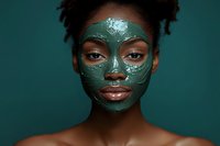 Young African American woman with green clay face mask for skincare routine and spa treatment on teal background.