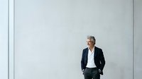 Senior male professional with gray hair and glasses wearing dark blazer and white shirt standing against white wall with hands in pockets, suitable for corporate business and leadership concepts.