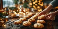 Freshly baked cookies on cooling rack with female hands arranging them, warm holiday bokeh lights in background creating cozy festive atmosphere.