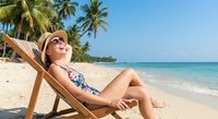 Young woman relaxing on beach chair wearing sunglasses and swimsuit on tropical sandy beach with palm trees and turquoise ocean water for summer vacation.