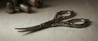 Ornate vintage scissors with decorative handles lie on neutral fabric surface alongside sewing notions and thimbles, perfect for crafting, tailoring, and needlework projects.