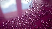 Water droplets bead on a deep burgundy surface, creating a hydrophobic effect with selective focus on glistening spheres against blurred background for product protection concepts.
