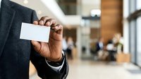Man in dark business suit holding blank white business card in modern office lobby with blurred background, suitable for corporate branding and professional networking concepts.