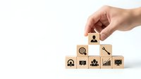 Hand placing wooden cube with business person icon on pyramid of blocks with corporate symbols for team building and leadership development.