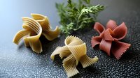 Raw pasta in shape of decorative flowers with fresh herbs on dark textured background. Colorful Italian farfalle and gigli pasta varieties create artistic culinary composition.