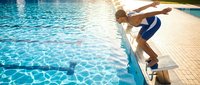 Athletic young man in blue swim shorts diving into crystal clear swimming pool from poolside deck during sunny summer day for recreation and fitness.