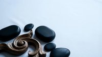 Young woman with long dark hair, wearing a white spa robe, receives a hot stone massage treatment on her back. Smooth black basalt stones arranged on skin for therapeutic relaxation and wellness.