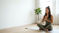Young woman with dark hair and medium skin tone practicing yoga or meditation on mat in bright, minimalist room with potted plant. Suitable for wellness, mindfulness, fitness, and lifestyle content.