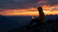 Woman working on laptop at mountain summit during golden sunset, silhouetted against dramatic sky. Remote work and digital nomad lifestyle concept.