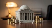 Classical bank building model surrounded by golden coin stacks on wooden desk with warm lamp lighting representing financial institution concept.