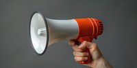 Hand holding red and white megaphone against gray background, demonstrating announcement or public speaking concept for business communication and marketing.