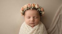 Peaceful newborn baby girl wearing delicate floral crown with soft pink roses sleeping wrapped in cream knit blanket for portrait photography.