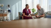 Female healthcare worker in burgundy scrubs consulting with elderly Caucasian male patient in bright hospital room with natural lighting and modern medical equipment.