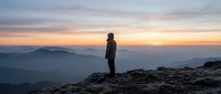 Silhouetted male hiker standing on rocky mountain peak at sunrise, gazing at misty valleys below. Outdoor adventure and nature exploration concept.