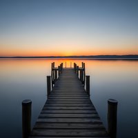 Wooden pier extending into calm lake at sunset with orange and blue sky reflection creating peaceful waterfront scene for relaxation and meditation concepts.
