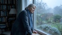 Mature man with gray hair and Caucasian ethnicity gazing thoughtfully out a window on a rainy day, wearing a cardigan in a library setting with bookshelves visible, contemplative mood for editorial and lifestyle use.