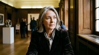 Professional mature Caucasian businesswoman in black suit standing confidently in elegant museum or gallery interior with wooden panels and artwork.