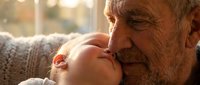 Elderly man with gray beard and wrinkles tenderly kissing infant's forehead by window. Grandfather and grandchild bonding moment showing intergenerational love and family connection.