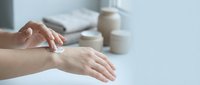 Woman applying white moisturizing cream on hands for skincare routine and hydration at home with cosmetic products in background.