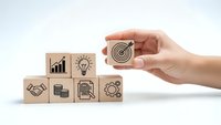 Hand holding wooden cube with target icon above business strategy blocks on white background for corporate planning and goal setting concepts.