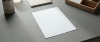 Clean minimalist workspace with blank white paper sheet on gray desk surrounded by modern office supplies and wooden organizer for professional presentations.