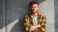 Confident middle-aged Caucasian man in yellow plaid shirt smiling with crossed arms against concrete wall in natural sunlight for lifestyle content.