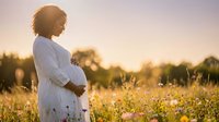 Pregnant African American woman in white dress standing in wildflower meadow at golden hour sunset, maternity lifestyle portrait for wellness content.