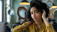 Young African American woman with natural curly hair wearing mustard yellow shirt looking pensive and concerned while sitting in modern cafe with vintage lighting fixtures.