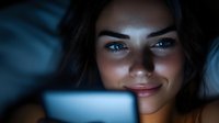 Young woman with dark hair and blue eyes, appearing to be of mixed ethnicity, smiling while looking at a glowing screen in a dark environment, suitable for technology, digital communication, and lifestyle content.