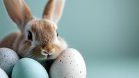 Adorable light brown bunny with long ears peeking behind pastel blue and speckled Easter eggs against soft turquoise background, creating festive spring atmosphere.