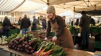 Young woman vendor in beige knit hat and brown apron arranging fresh organic vegetables at bustling farmers market stall with striped canopy tent.