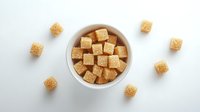Brown sugar cubes in white ceramic bowl on white background, scattered pieces around. Clean minimal food composition for cooking and baking projects.
