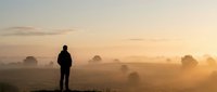 Man silhouette standing in misty field during golden sunrise with warm light rays and peaceful countryside landscape for meditation and inspiration concepts.