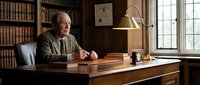 Mature man with gray hair and glasses, wearing a brown jacket, sitting at wooden desk in traditional library office with bookshelves, brass lamp, and window light for professional, academic, or business concepts.