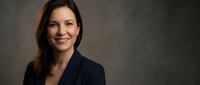 Professional businesswoman with dark hair smiling confidently in navy blazer against neutral background for corporate headshots and business presentations.